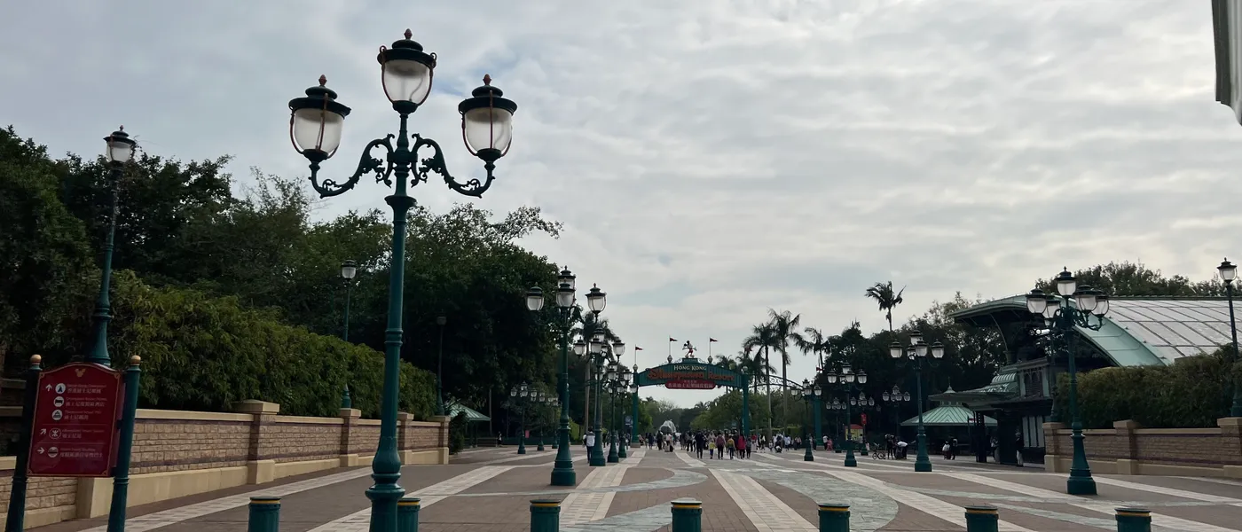 A quiet, near-empty entrance walkway at Hong Kong Disneyland lined with lamp posts and palm trees under an overcast sky